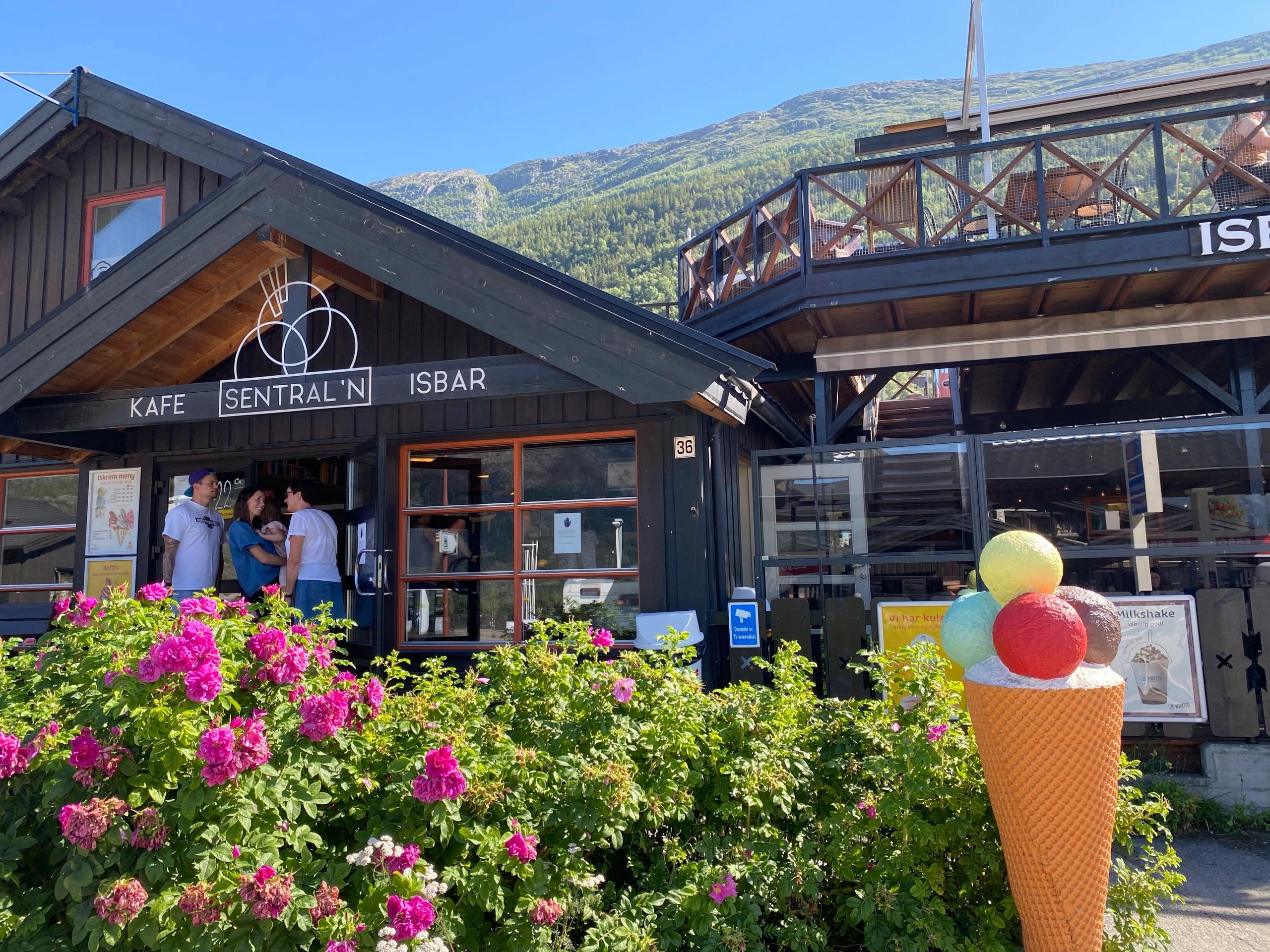 Mørk trebygning med skiltet «KAFE SENTRAL’N ISBAR», rosa blomar i forgrunnen og fjell i bakgrunnen..
