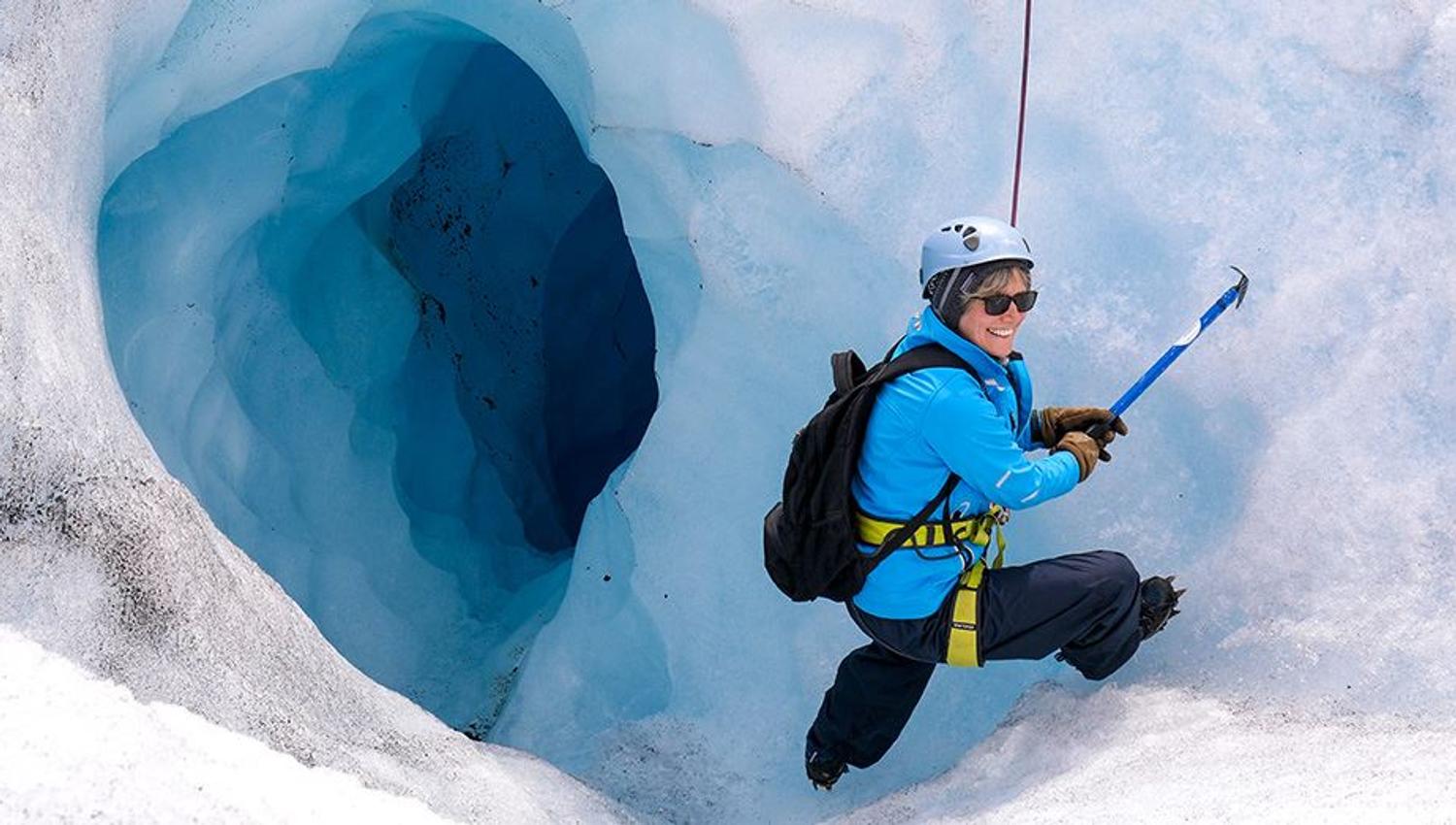 A climber wearing helmet and backpack uses an ice axe on a snowy glacier beside a deep blue crevasse opening..