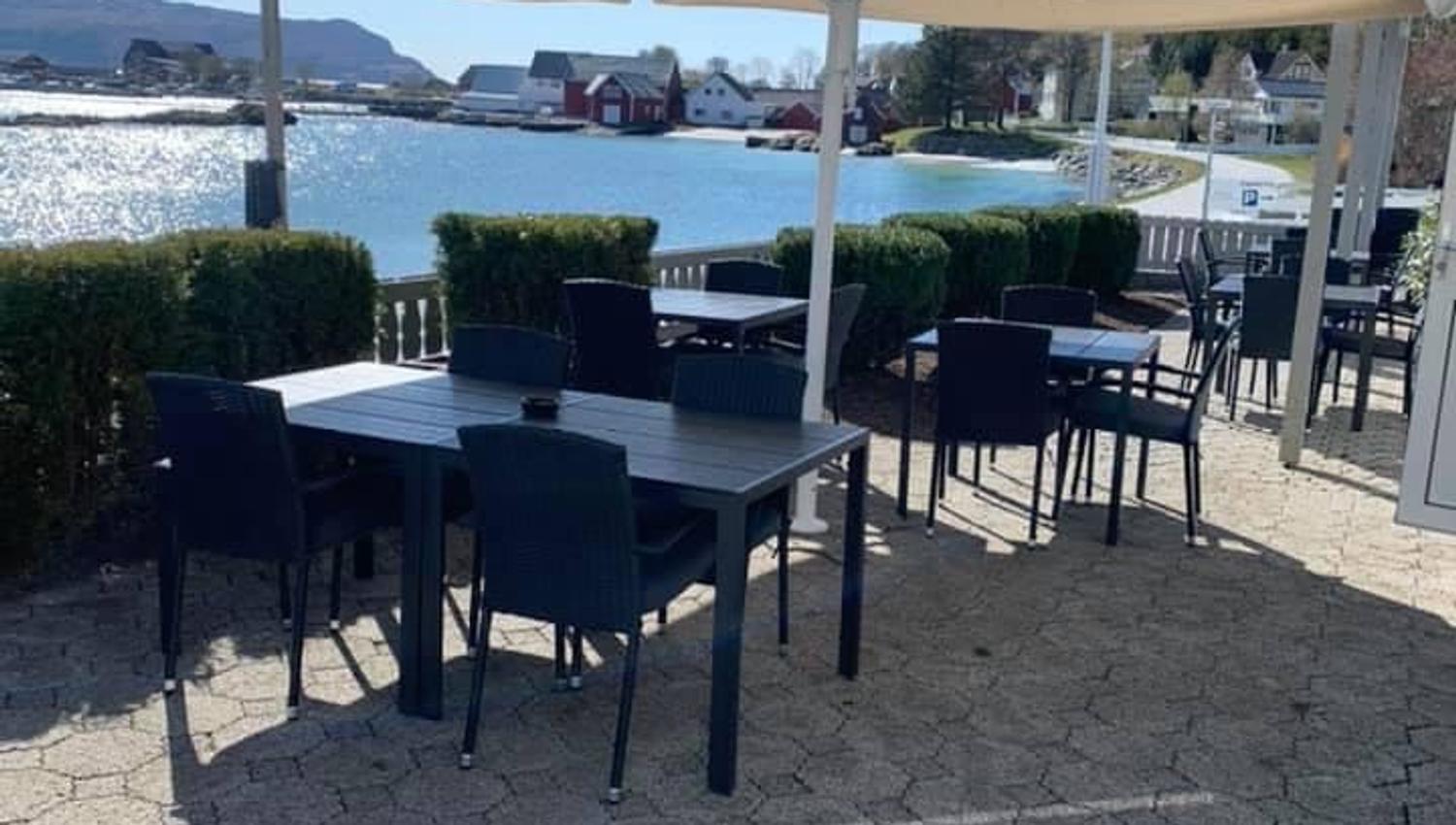 Outdoor patio with black tables and chairs under a large umbrella beside a calm waterfront and distant houses..