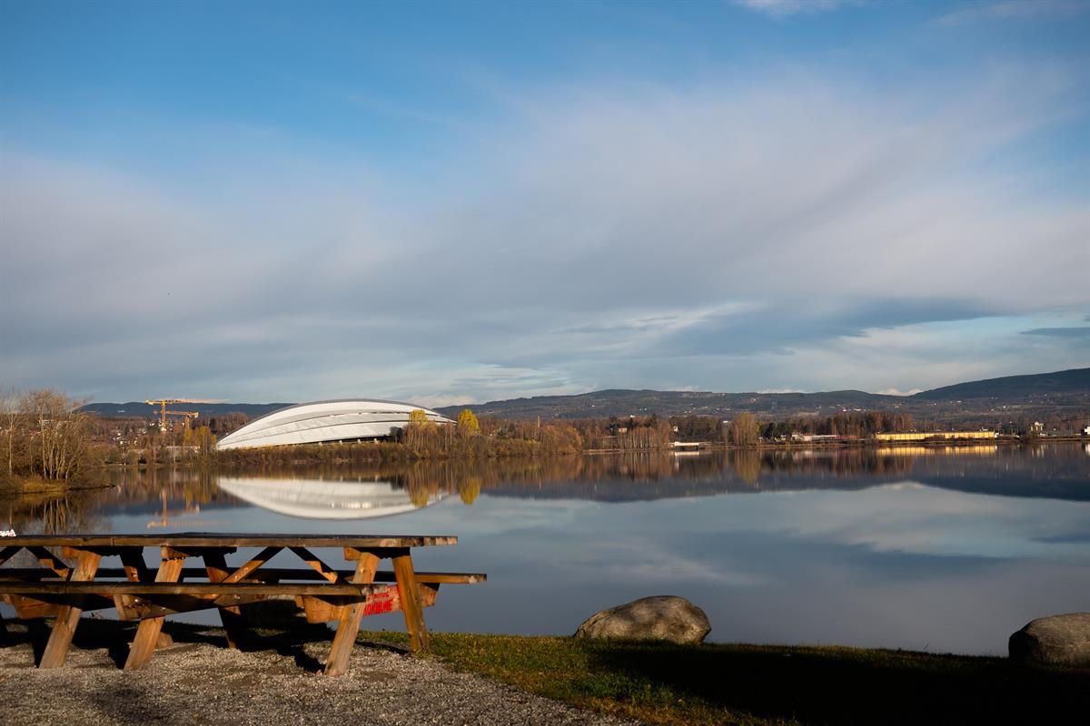 Outdoor area by the water with picnic benches in the foreground and a building with a curved roof across the water. Calm autumn landscape surrounds th