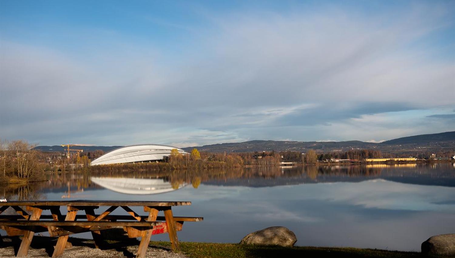 Outdoor area by the water with picnic benches in the foreground and a building with a curved roof across the water. Calm autumn landscape surrounds th