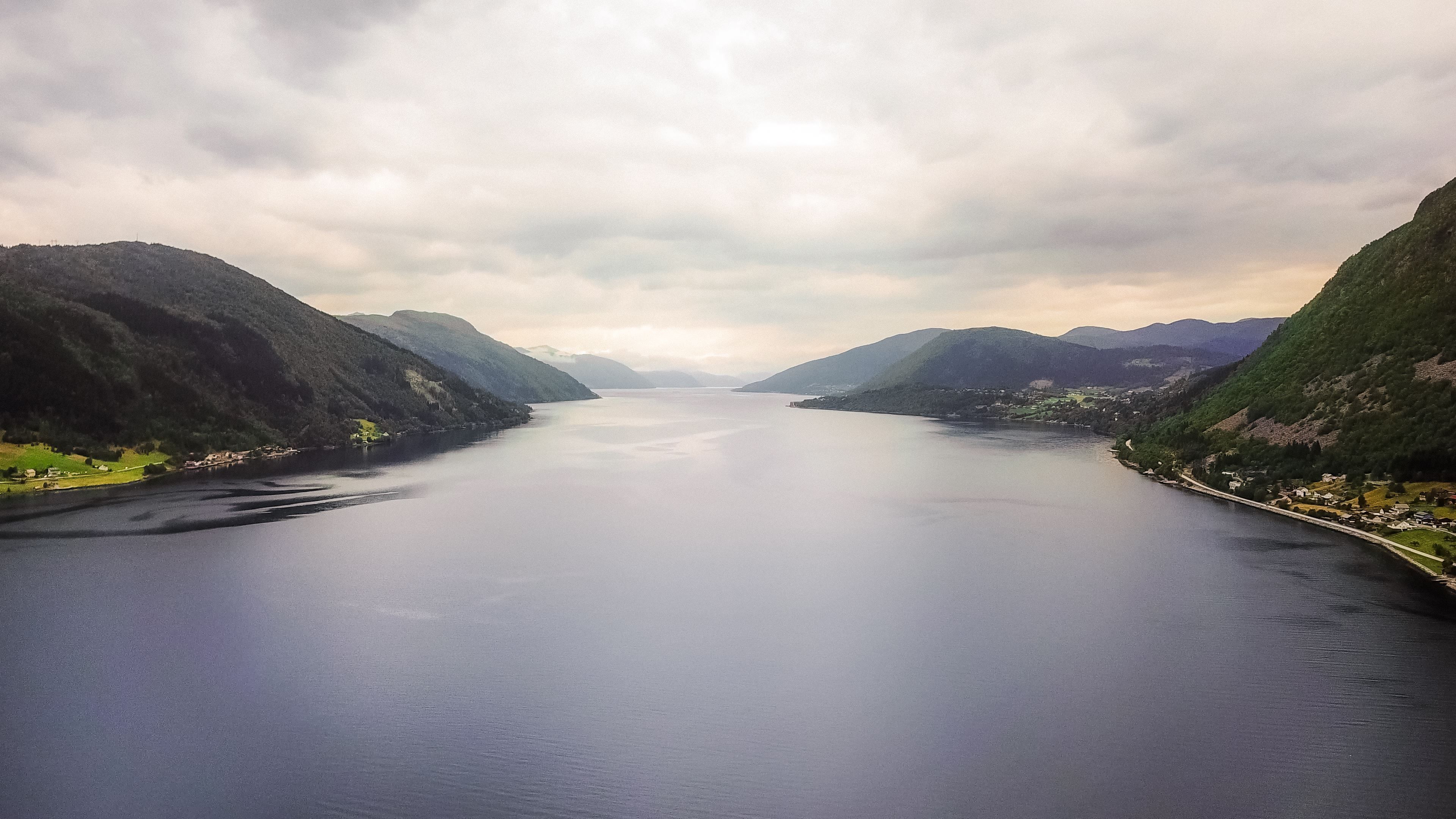 Wide view of a calm fjord between green mountains under an overcast sky, with small shoreline settlements and a road.