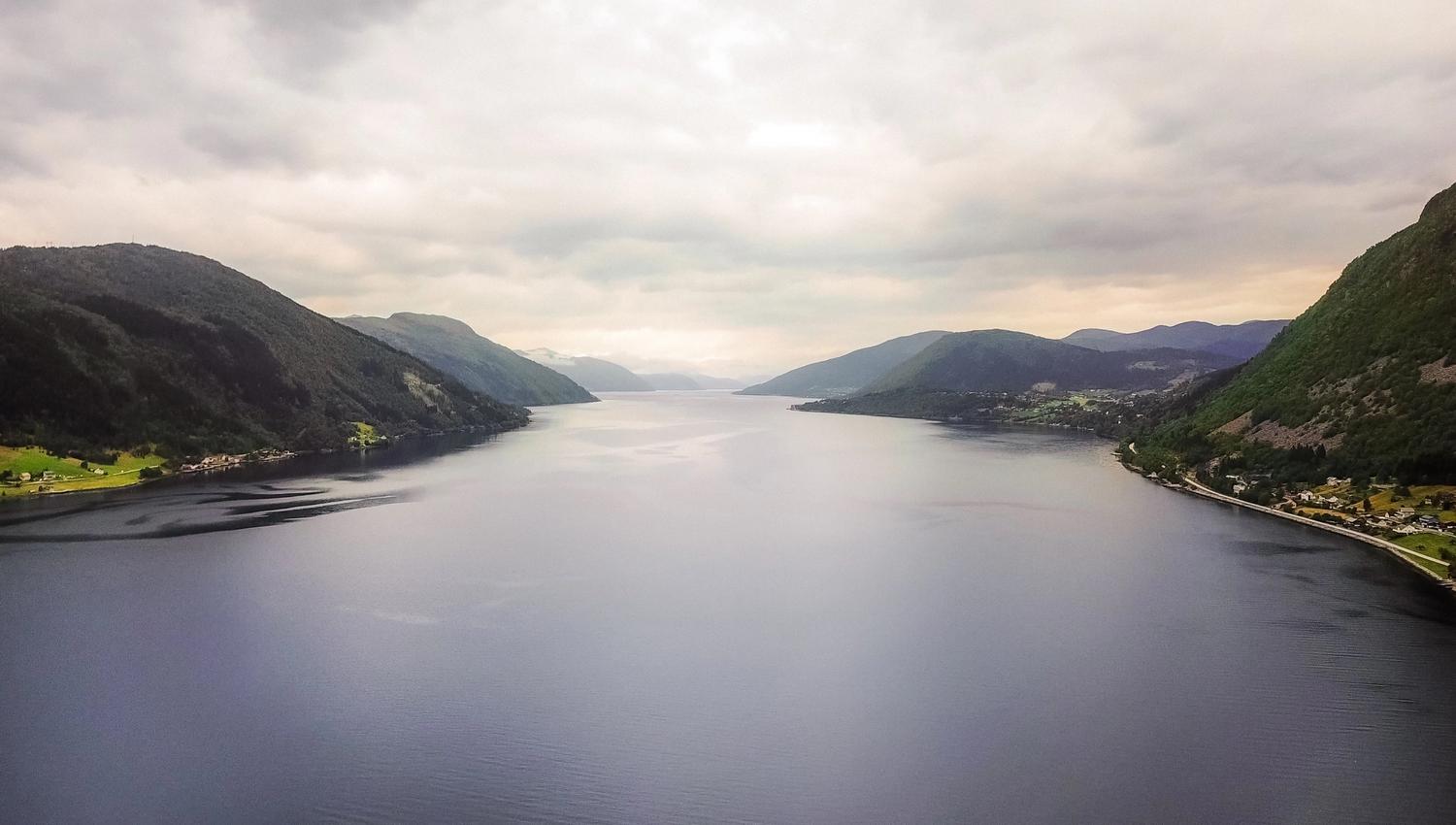 Wide view of a calm fjord between green mountains under an overcast sky, with small shoreline settlements and a road.