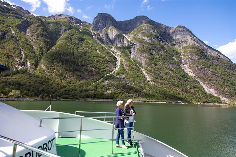 To passasjerar står ved rekkverket i baugen på fjordcruisebåten og ser mot bratte fjellsider ved Hardangerfjorden.