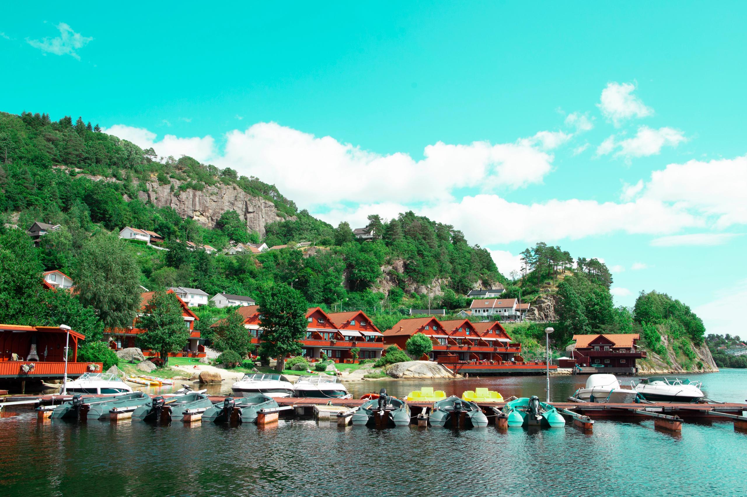 Red-brown holiday cabins by the water, and a pier filled with boats. Forest and mountains rise behind the cabins, under a blue sky with a few scattere