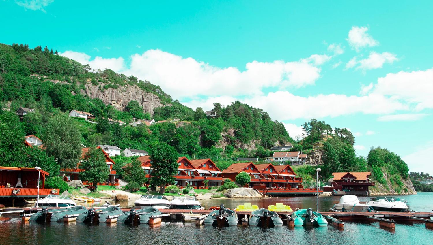 Red-brown holiday cabins by the water, and a pier filled with boats. Forest and mountains rise behind the cabins, under a blue sky with a few scattere