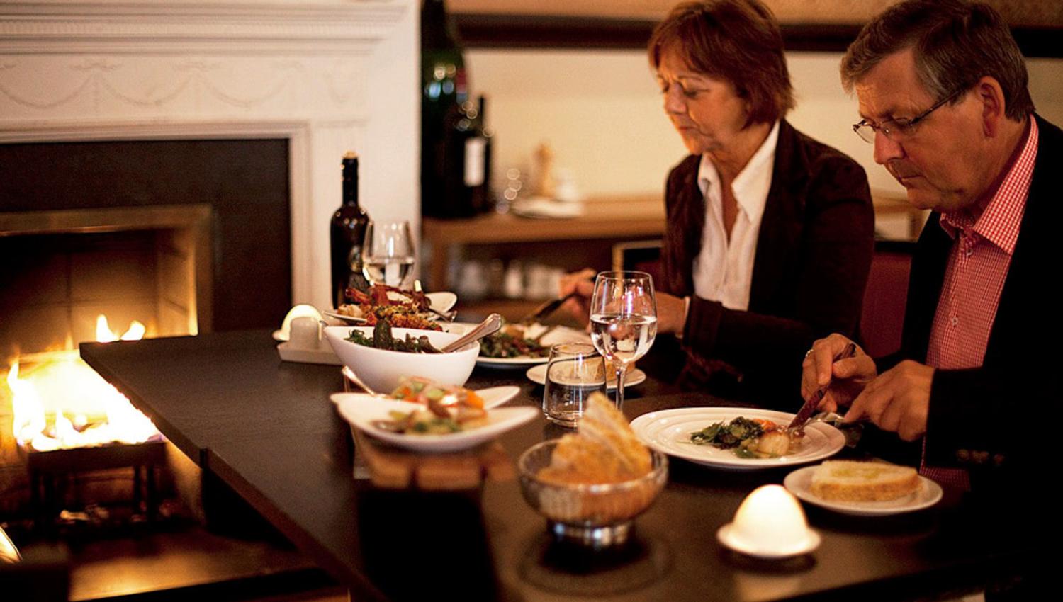 A man and a woman eating dinner next to a fireplace.
