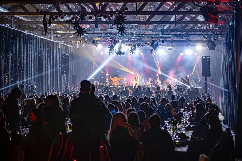 Large audience seated at banquet tables watching performers on a lit stage with beams, haze, and a disco ball overhead.