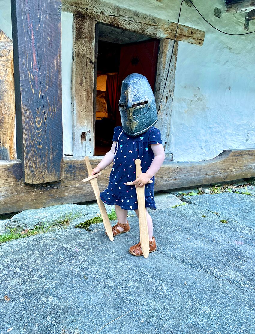 A child with a medieval helmet and two wooden swords is posing for a picture. 