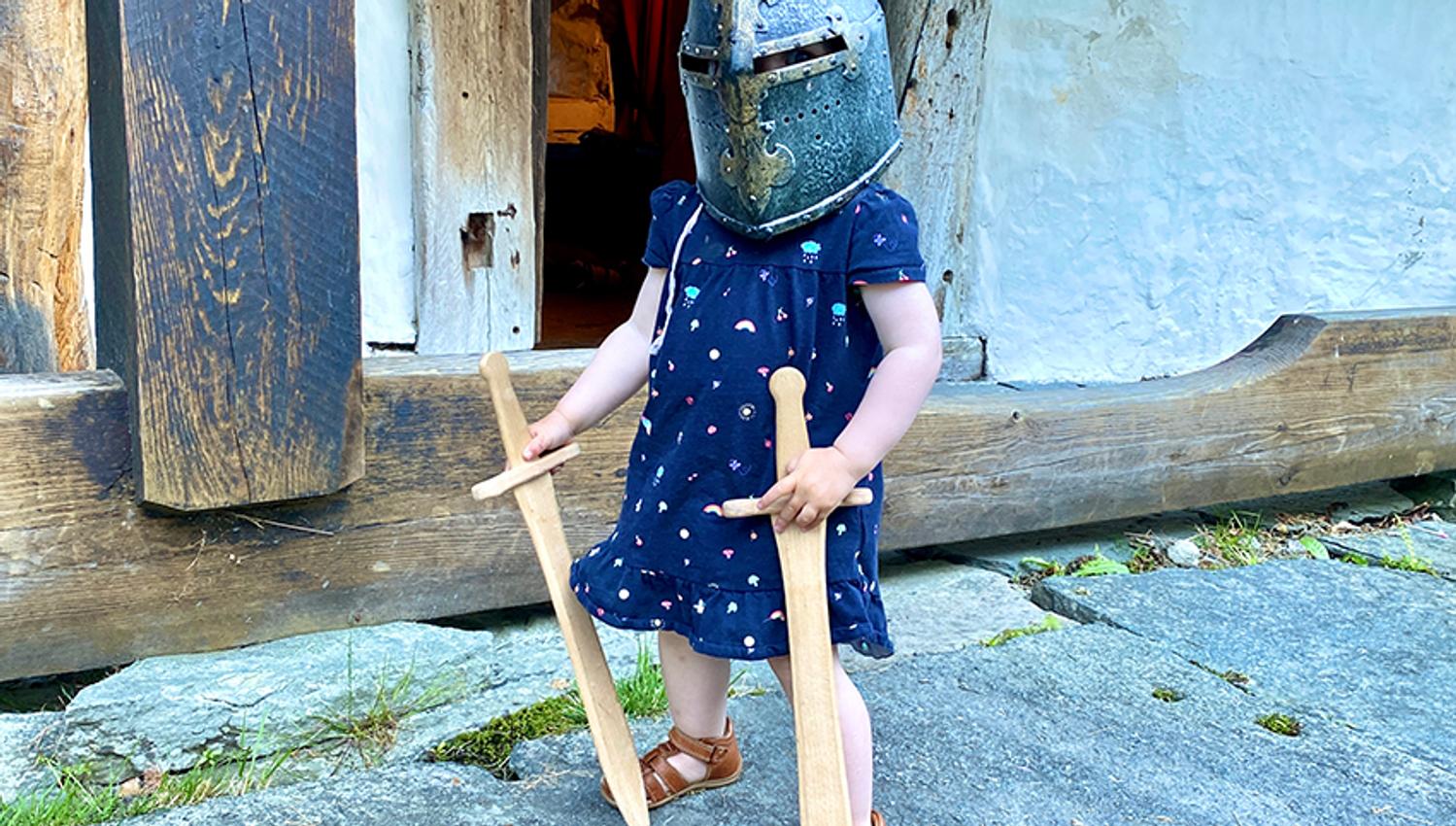 A child with a medieval helmet and two wooden swords is posing for a picture.
