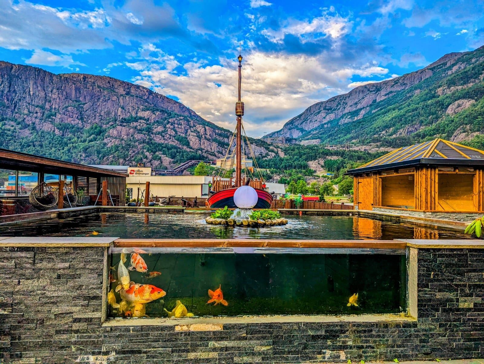 Utandørs fiskedam med synlege koi i glasfront, trebygningar og fjell i bakgrunnen under blå himmel..