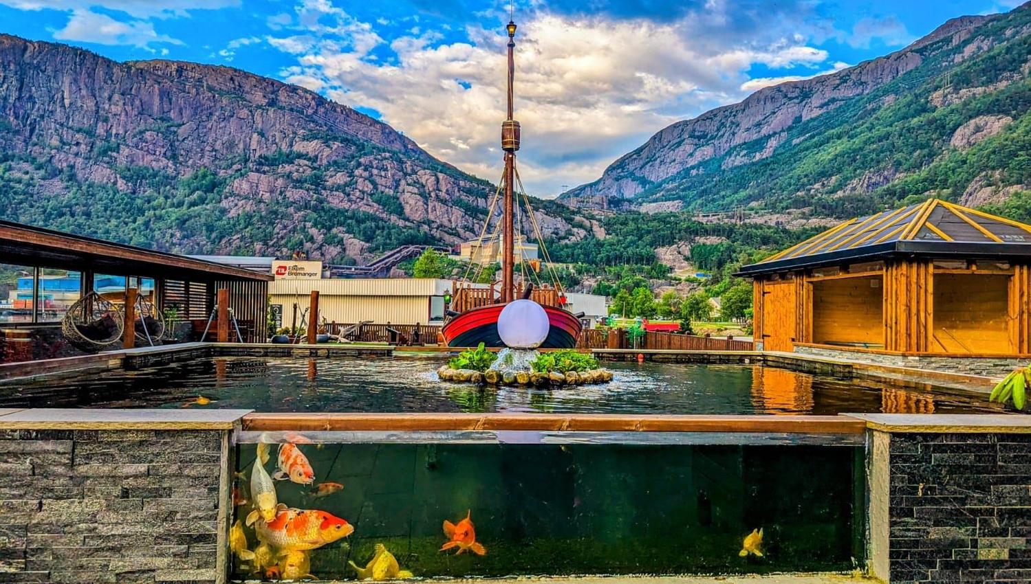 Utandørs fiskedam med synlege koi i glasfront, trebygningar og fjell i bakgrunnen under blå himmel..