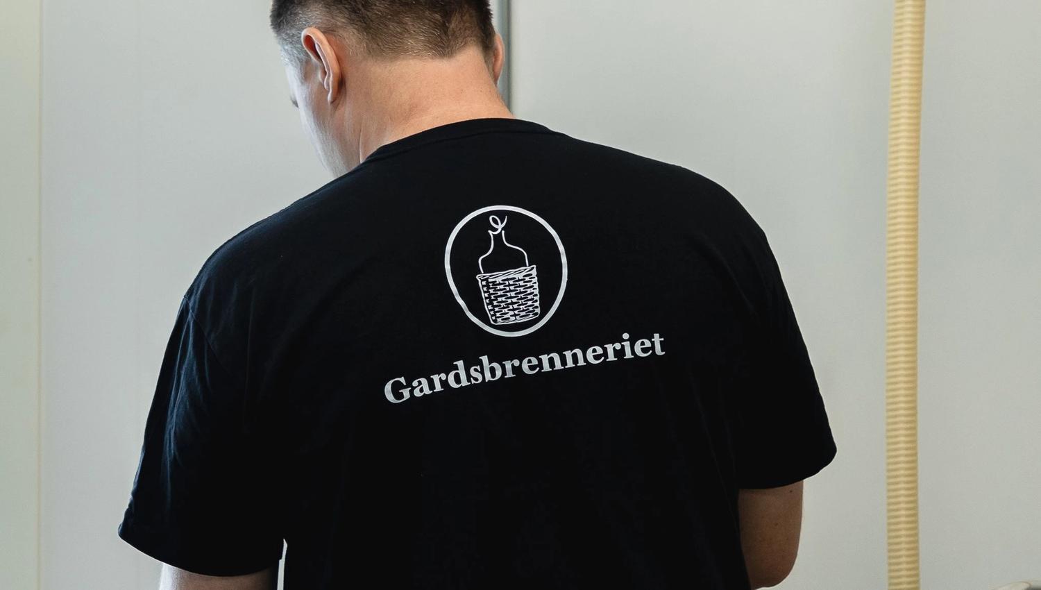 Rear view of a person wearing a black T-shirt with a white logo and the text “Gardsbrenneriet” indoors.