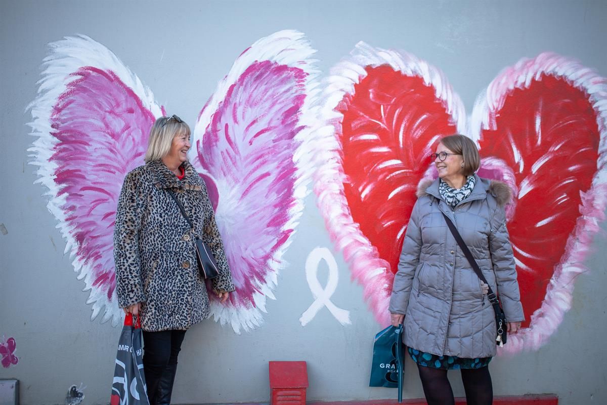 Two persons are posing in front of a wall decorated with large, colorful wing illustration, a popular photo spot associated with shopping in the city 