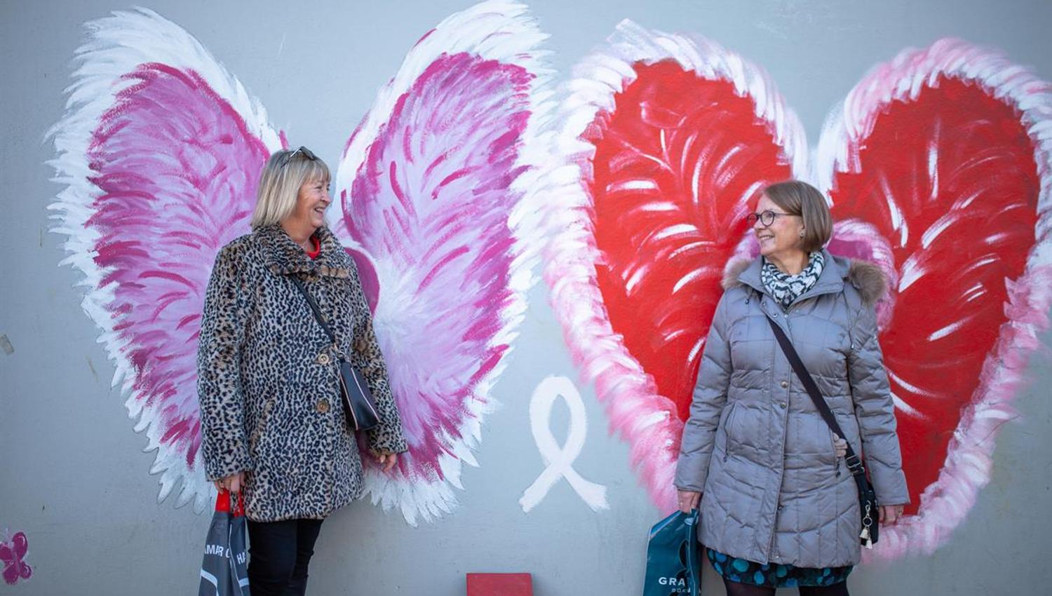 Two persons are posing in front of a wall decorated with large, colorful wing illustration, a popular photo spot associated with shopping in the city