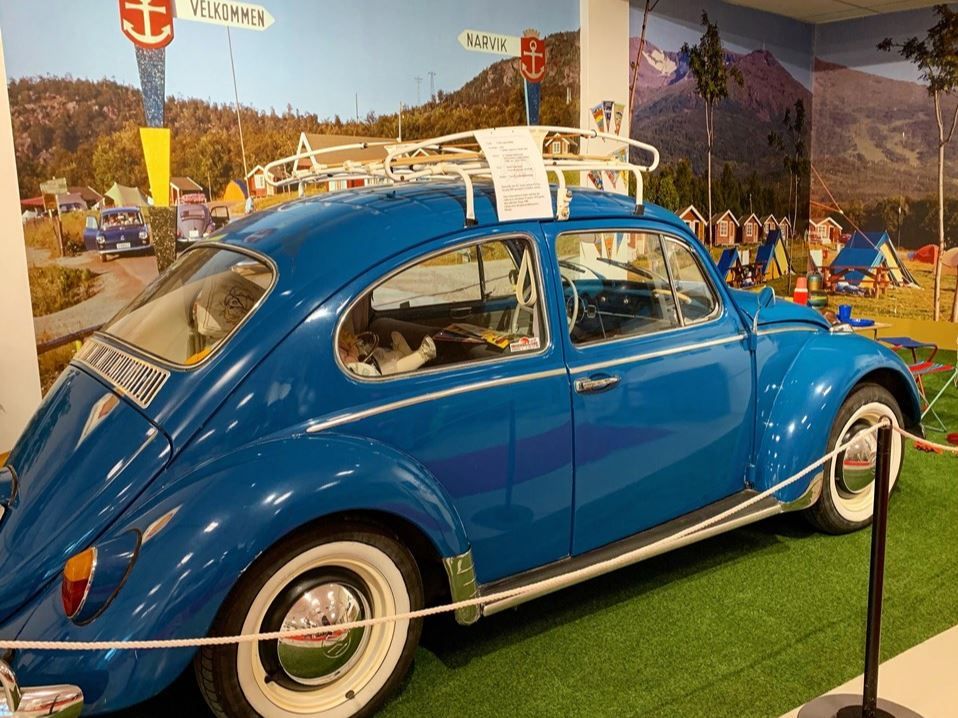 Blue vintage Volkswagen Beetle with roof rack displayed indoors behind a rope, with a scenic mural backdrop.