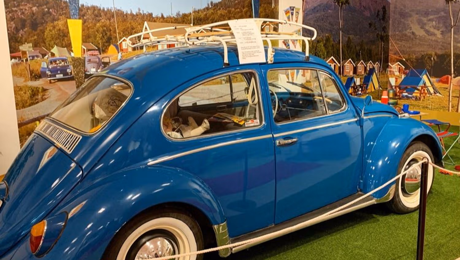 Blue vintage Volkswagen Beetle with roof rack displayed indoors behind a rope, with a scenic mural backdrop.