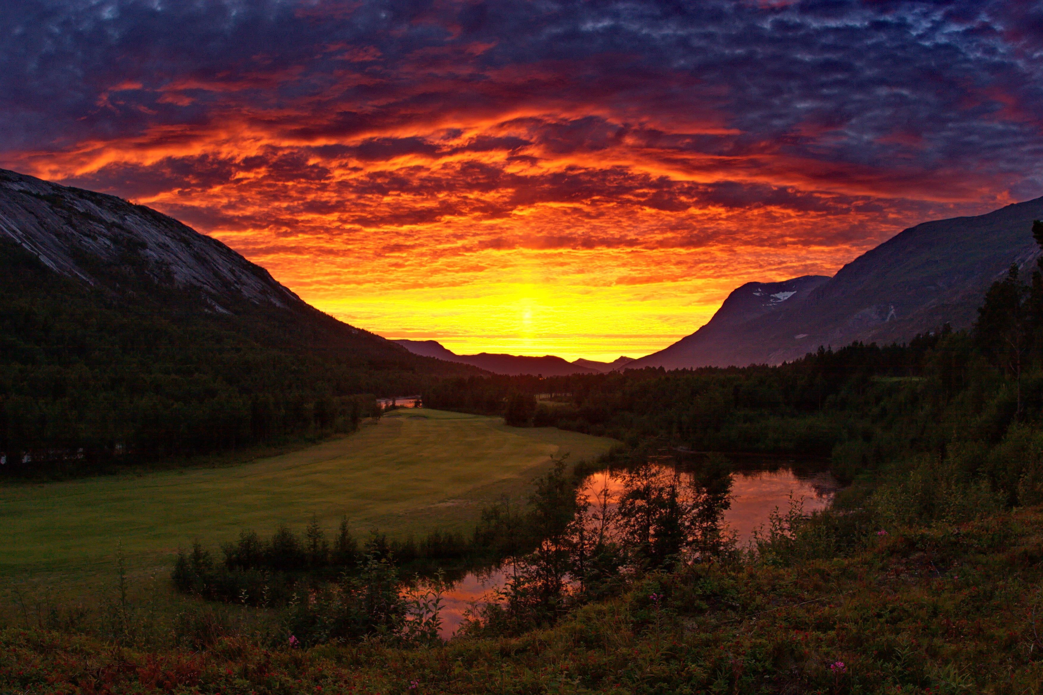 Brilliant orange sunset clouds above a mountain valley with forest, grassy meadow, and a river reflecting the sky..