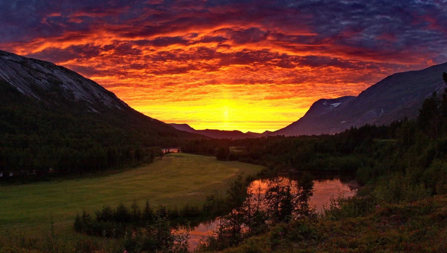 Brilliant orange sunset clouds above a mountain valley with forest, grassy meadow, and a river reflecting the sky..