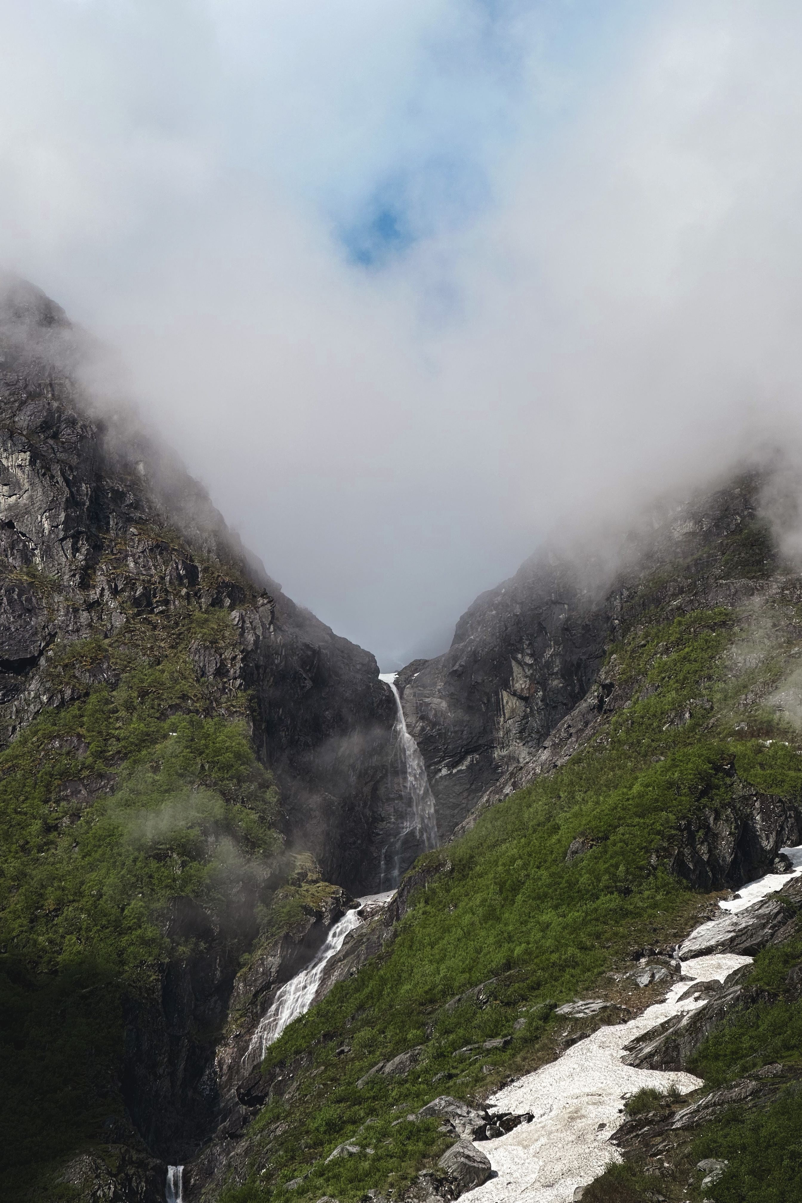 A tall waterfall drops between steep rocky green mountains, with low clouds and mist partially covering the peaks..