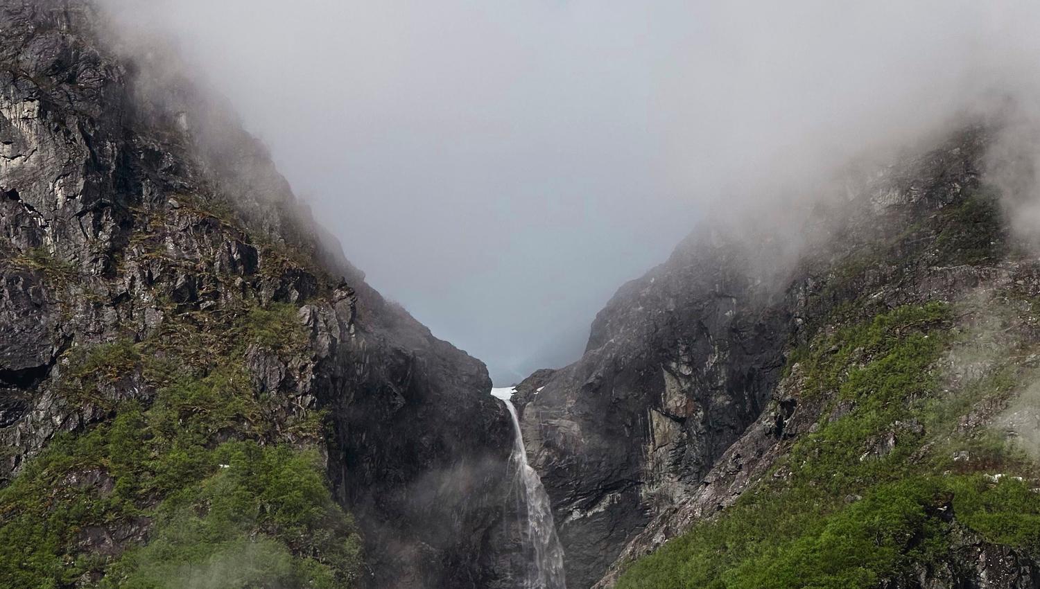 A tall waterfall drops between steep rocky green mountains, with low clouds and mist partially covering the peaks..