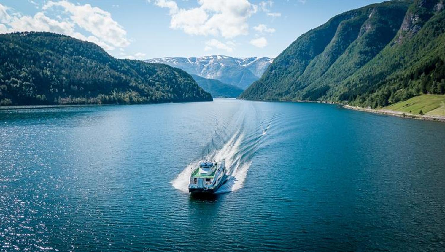 Fjordcruisebåten på Hardangerfjorden sett frå drone forfra, med blått vatn og snødekte fjell på begge sider.