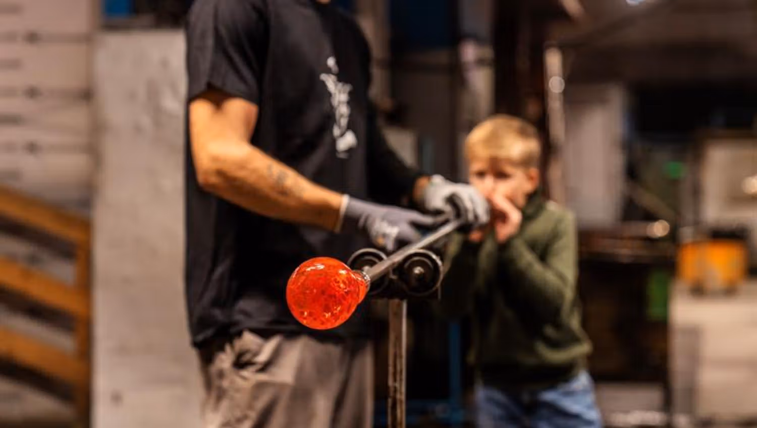 En gutt blåser glass med hjelp fra en glassblåser, mens den glødende glassmassen formes på røret