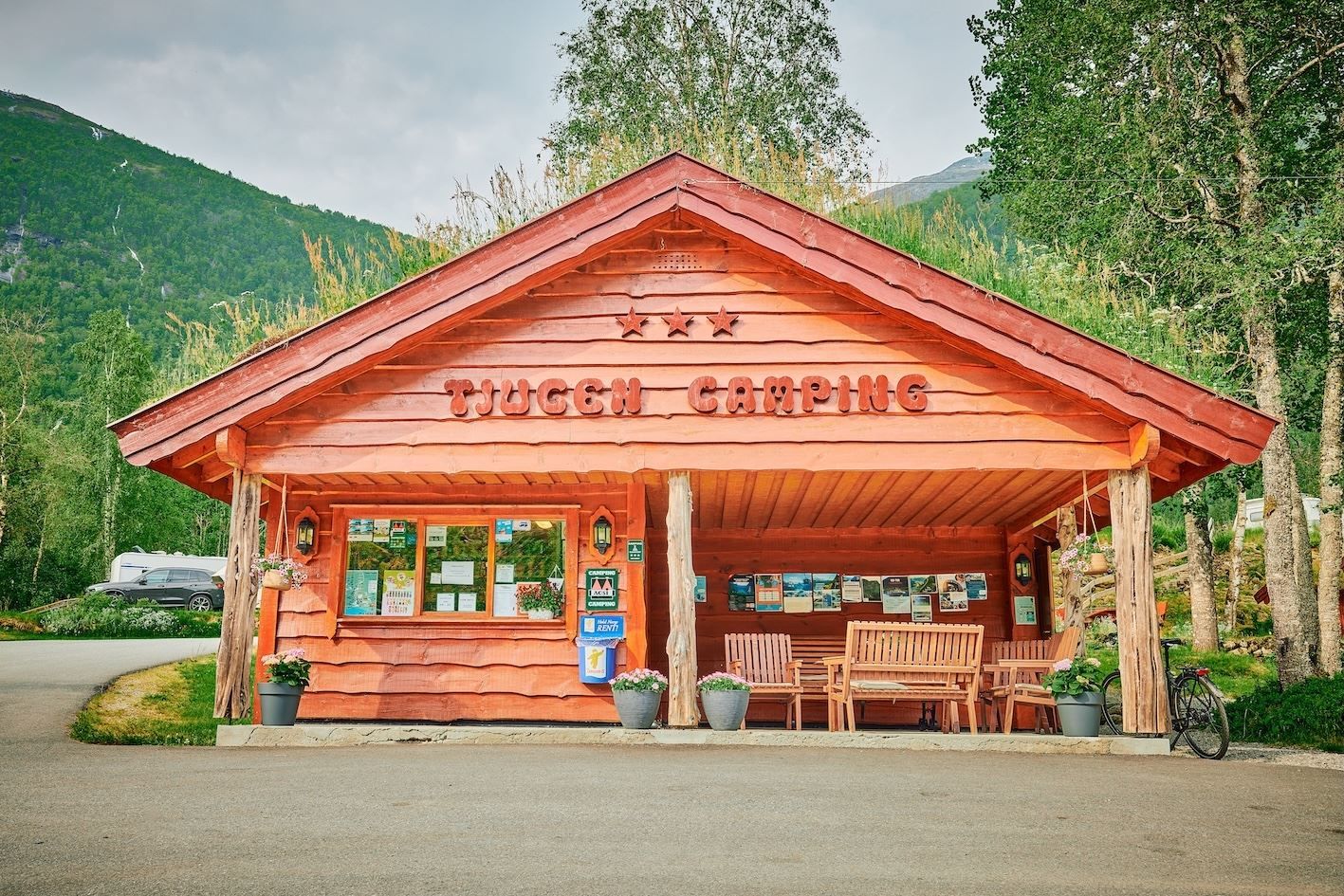 Raudt trebygg merka «Tjugum Camping» med benkar og plakatar under tak, omgitt av tre og fjell..