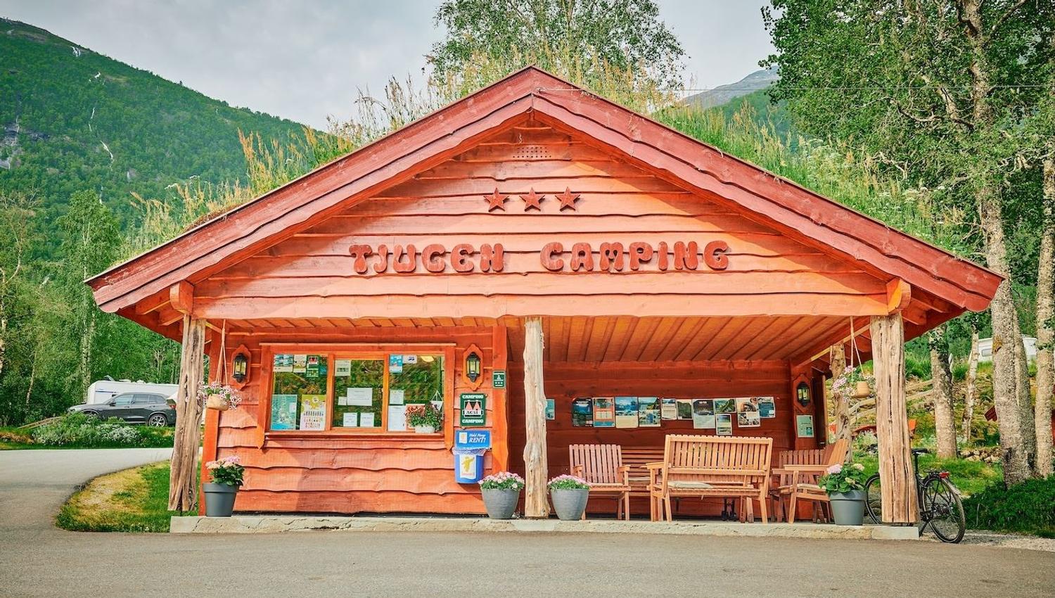 Raudt trebygg merka «Tjugum Camping» med benkar og plakatar under tak, omgitt av tre og fjell..