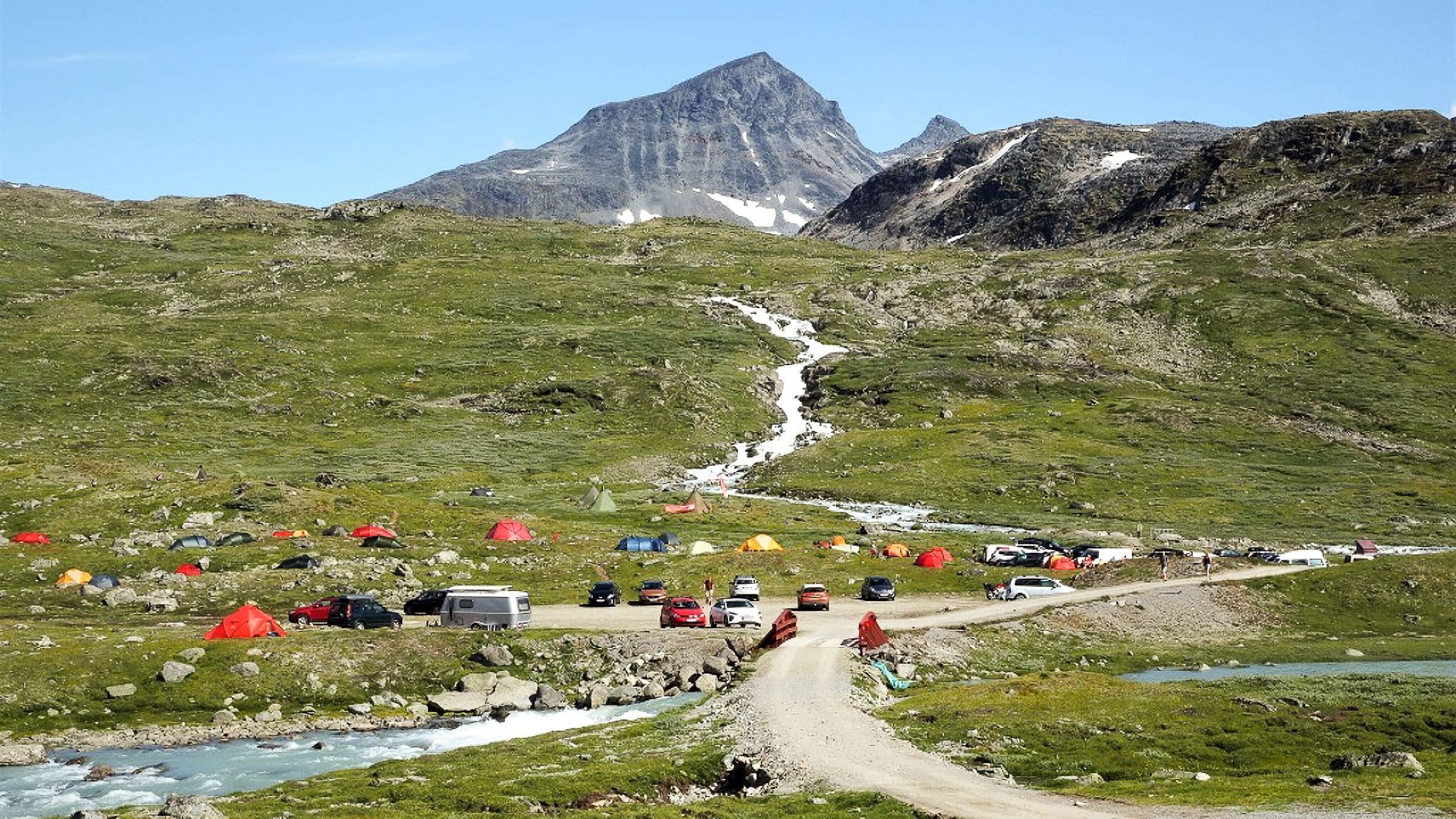A green alpine valley with colorful tents and parked cars near a dirt road and stream, below a rocky mountain peak..