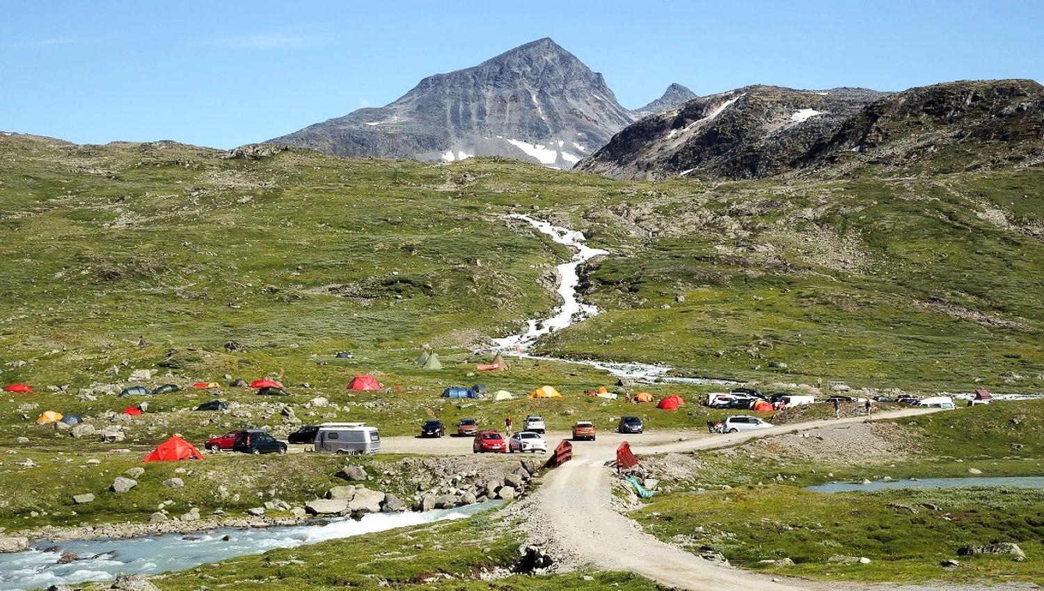 A green alpine valley with colorful tents and parked cars near a dirt road and stream, below a rocky mountain peak..
