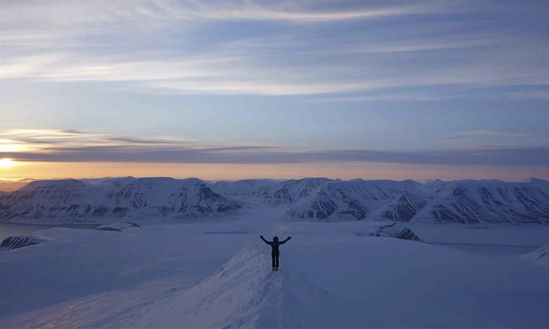 A person with outstretched arms on a snowy mountain ridge, with dark snow-covered mountains and a sunset in the background