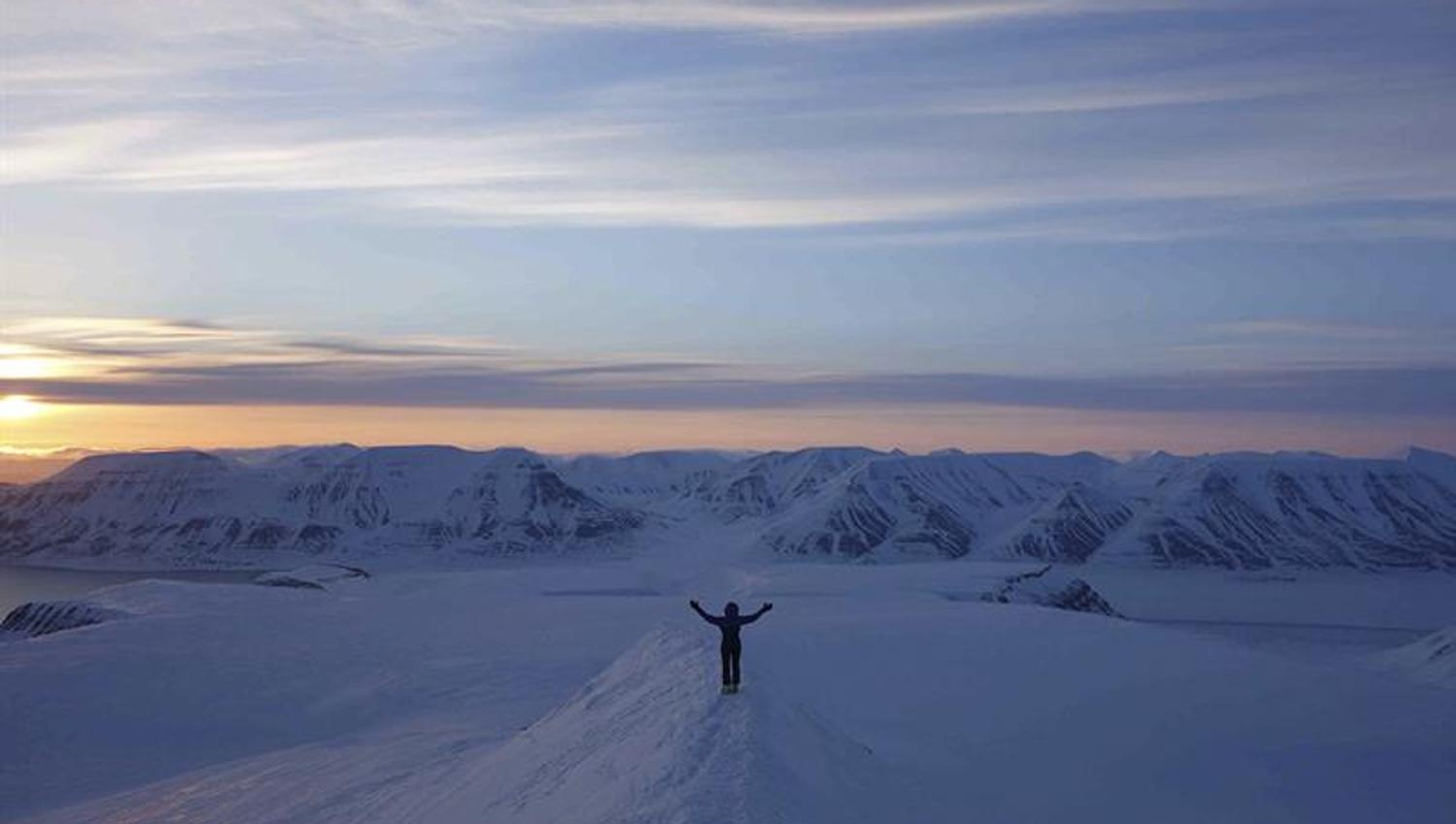 A person with outstretched arms on a snowy mountain ridge, with dark snow-covered mountains and a sunset in the background