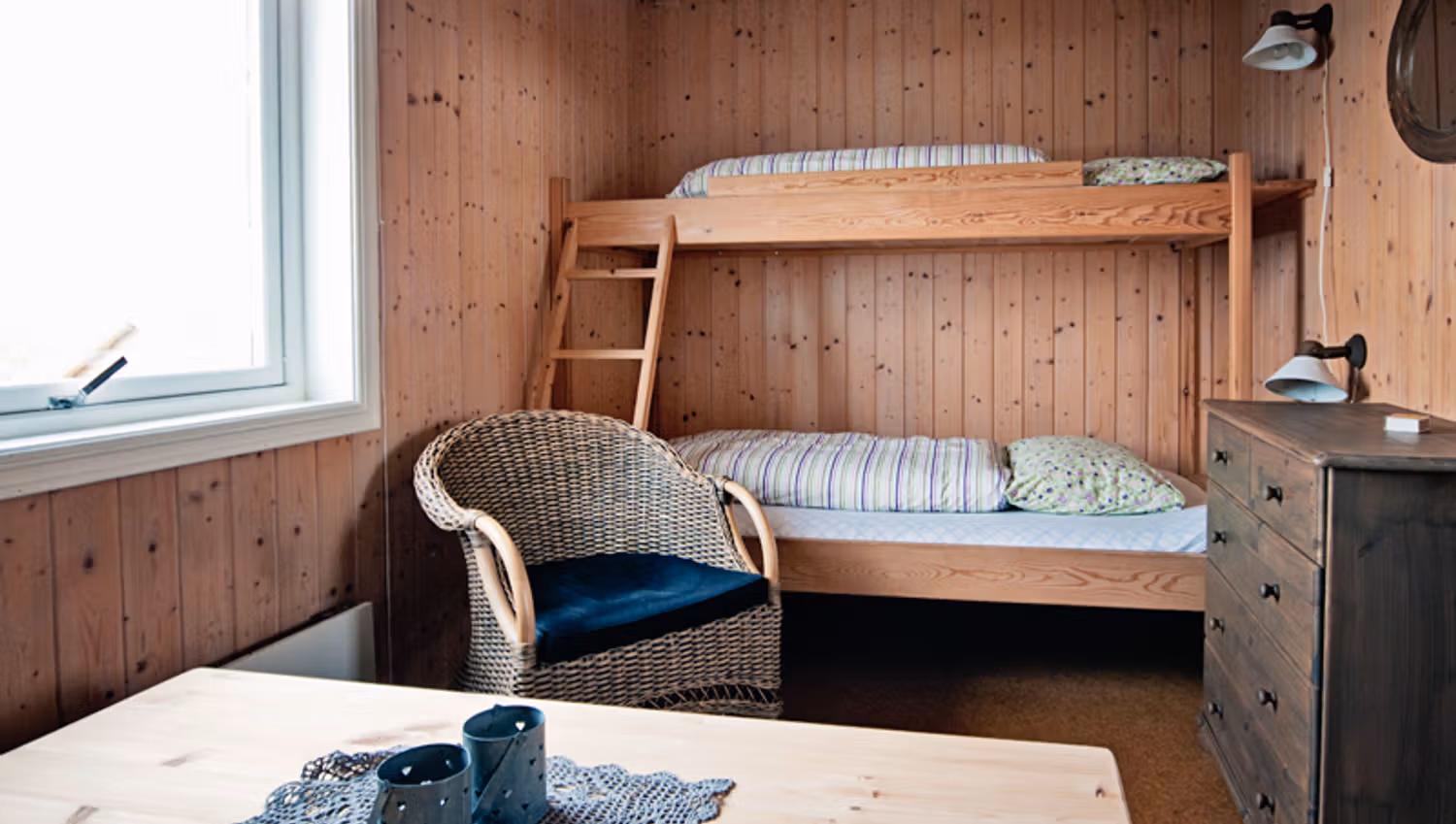 A room with wooden walls, a bunk bed, dresser, chair and table.