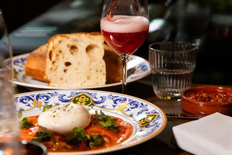 A bottle pouring a pink drink into a wine glass over a table with burrata on tomato, bread, small plates and a glass of water.