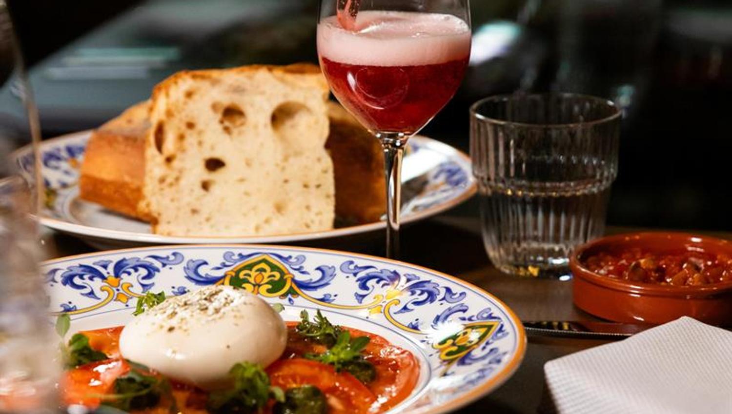 A bottle pouring a pink drink into a wine glass over a table with burrata on tomato, bread, small plates and a glass of water.