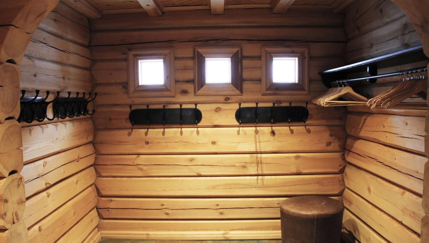 Interiör av en träpanelad hall med tre små fönster, väggkrokar, galgar på stång och en rund sittpuff.