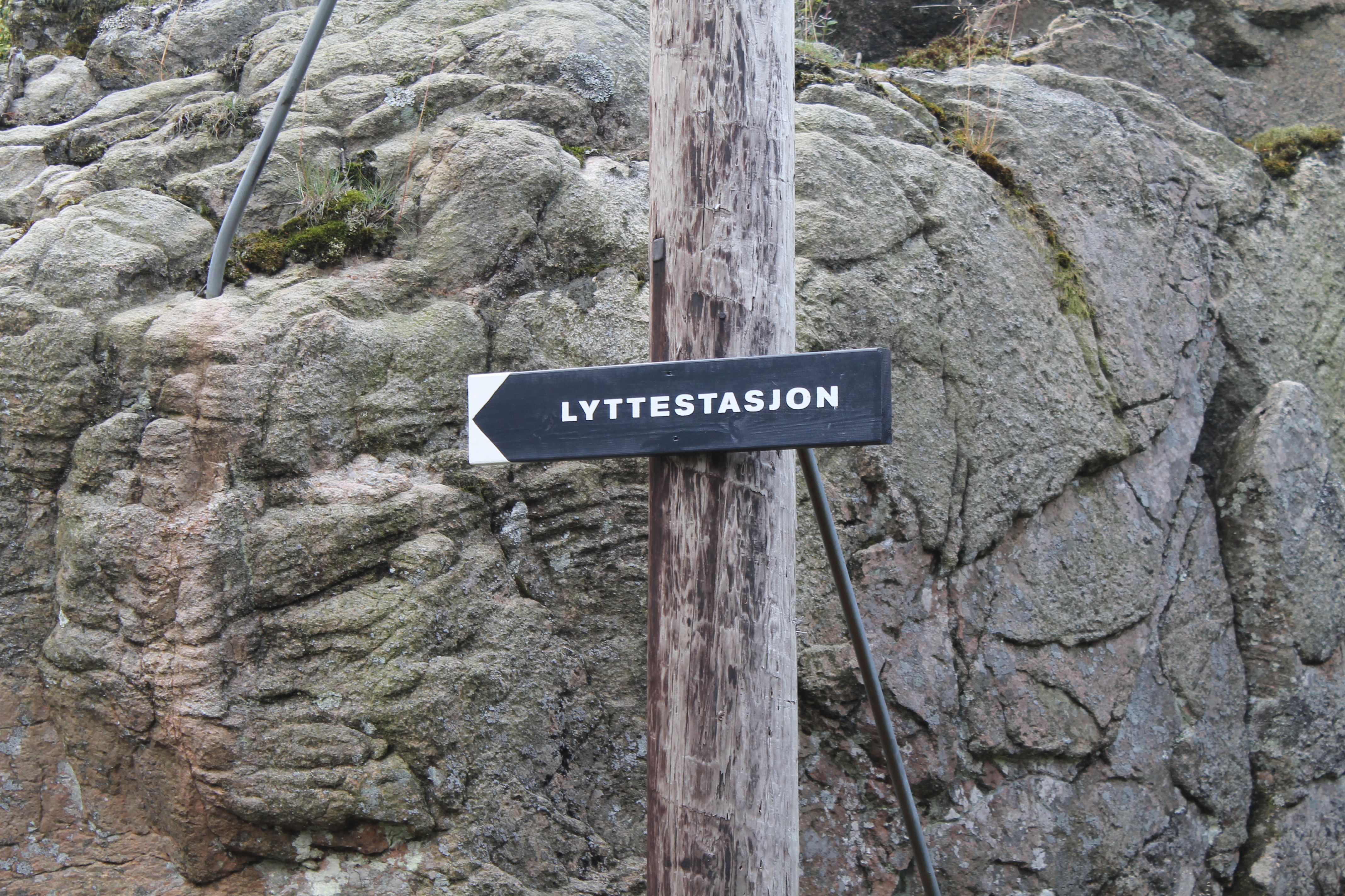 Svart skilt med hvit skrift "Lyttestasjon" på en stolpe
