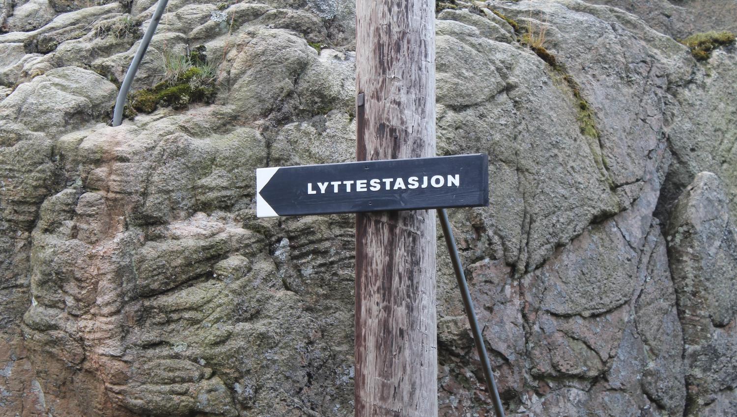 Svart skilt med hvit skrift "Lyttestasjon" på en stolpe