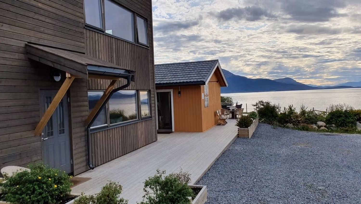 Wooden house by the water with mountains in the background and a garden in front