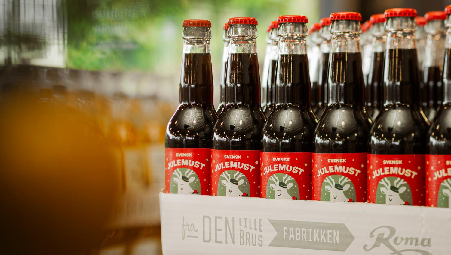 Brett med mørke glassflasker merket «Svensk Julemust» med røde etiketter, plassert på en butikkhylle.