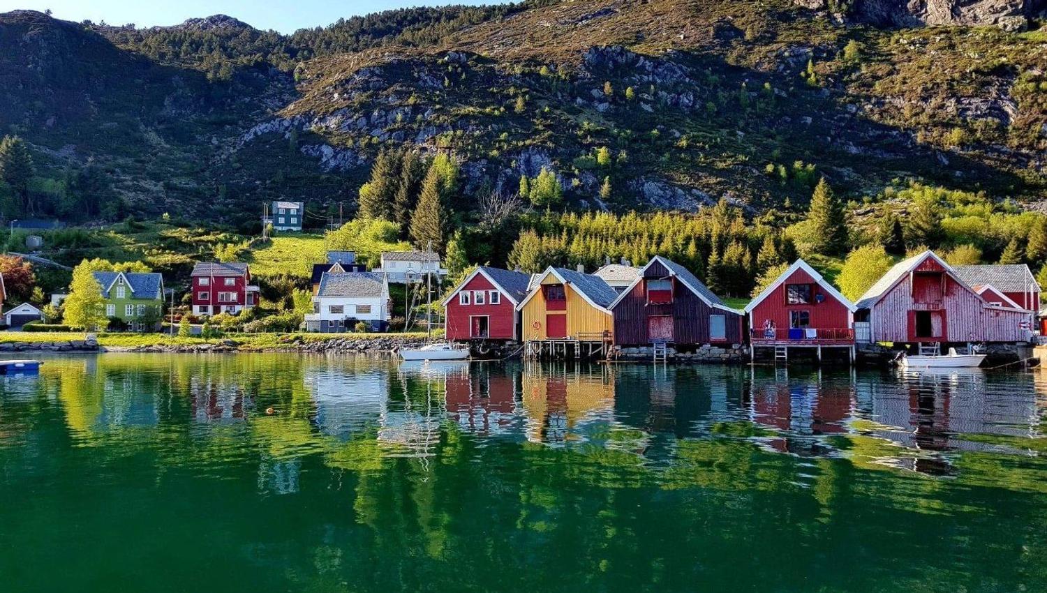 Red and yellow wooden boathouses line a calm fjord, reflecting in green water beneath rocky, tree-covered hills..