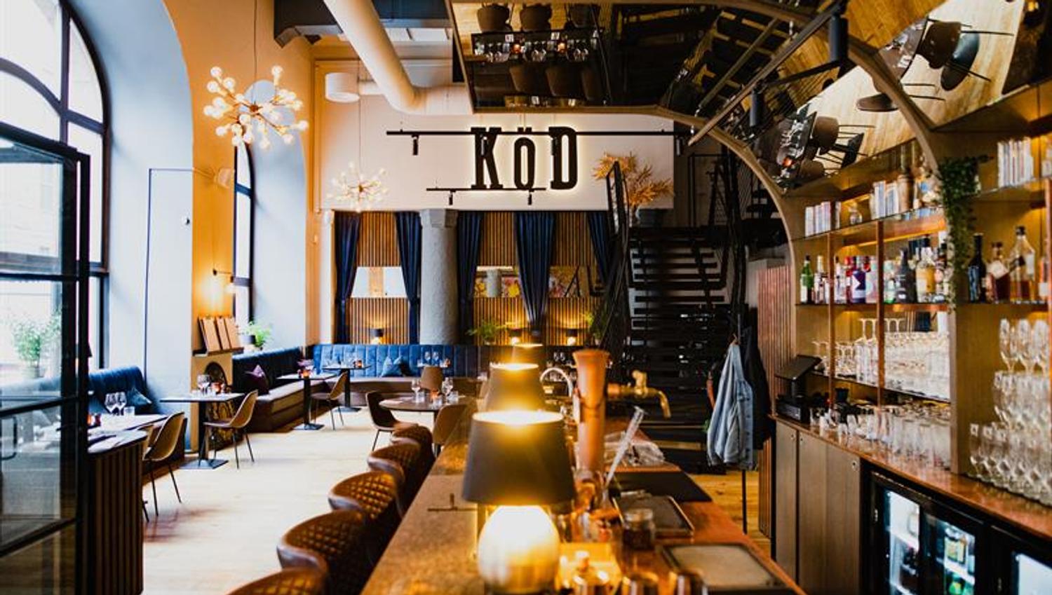 Restaurant interior with bar counter, bar stools, shelves with glasses and wine bottles, blue seating section along the wall and a high ceiling