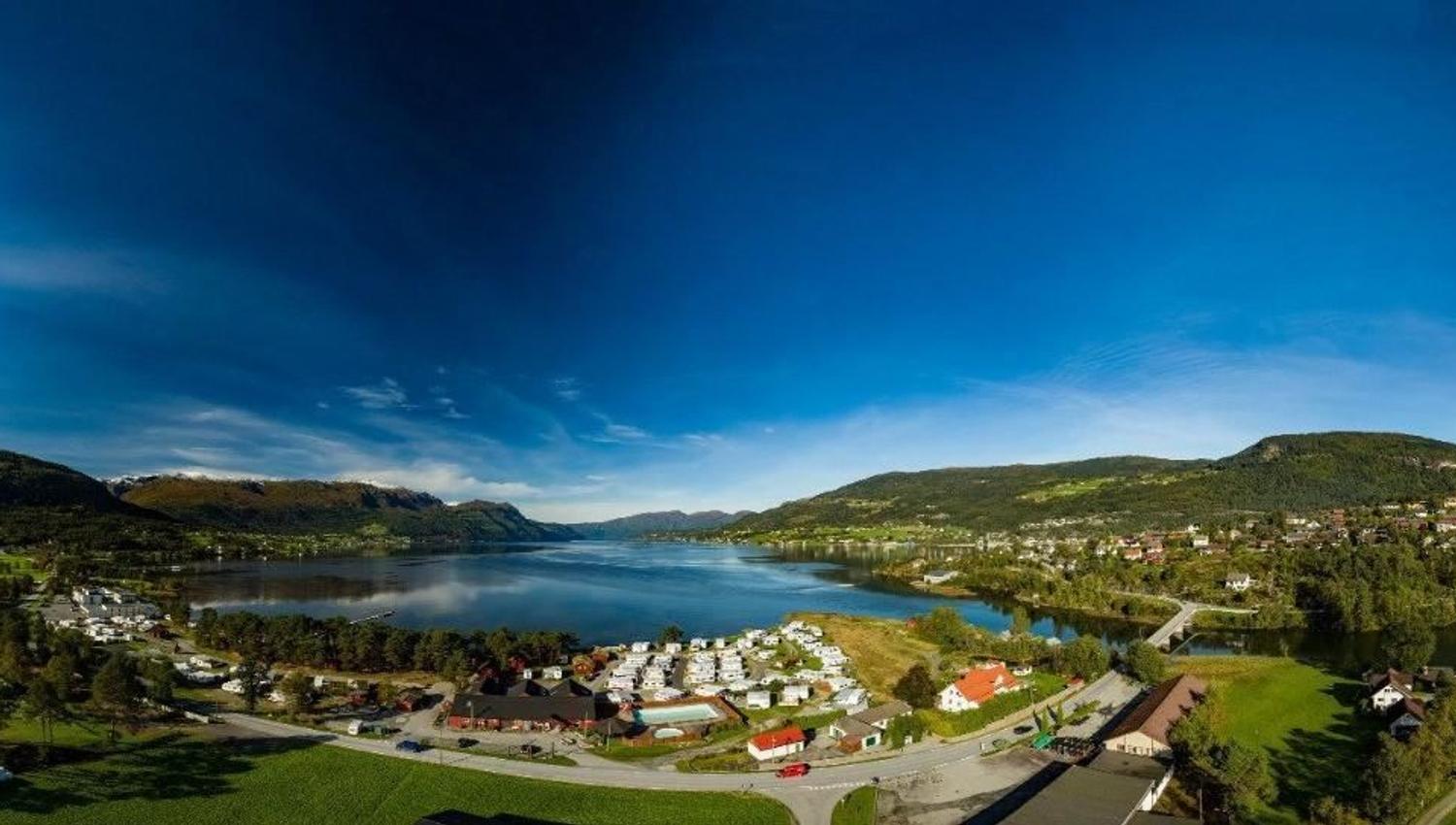 Wide panoramic view of Gloppen Camping in Sandane by Gloppenfjorden with green fields, scattered houses, roads, and mountains under a blue sky..