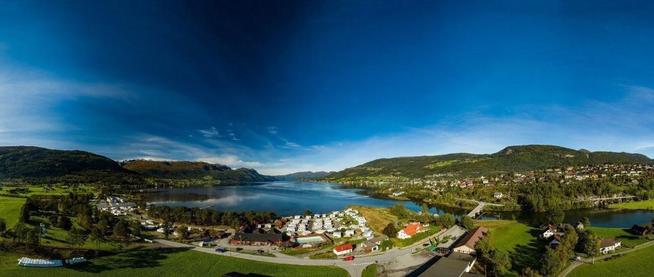 Wide panoramic view of Gloppen Camping in Sandane by Gloppenfjorden with green fields, scattered houses, roads, and mountains under a blue sky..