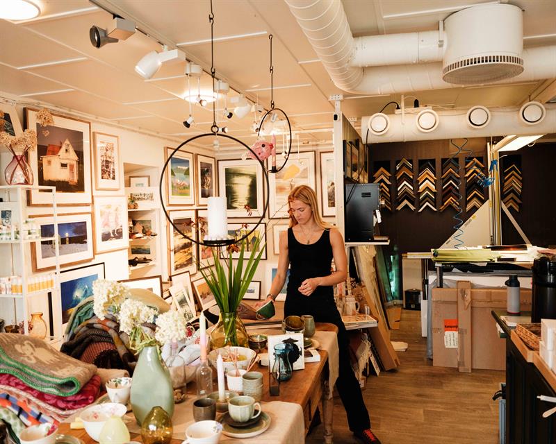 Inside the gallery at Iversen Bakery and Gallery, a woman works with ceramics and interior items surrounded by artworks, frames and handmade products.