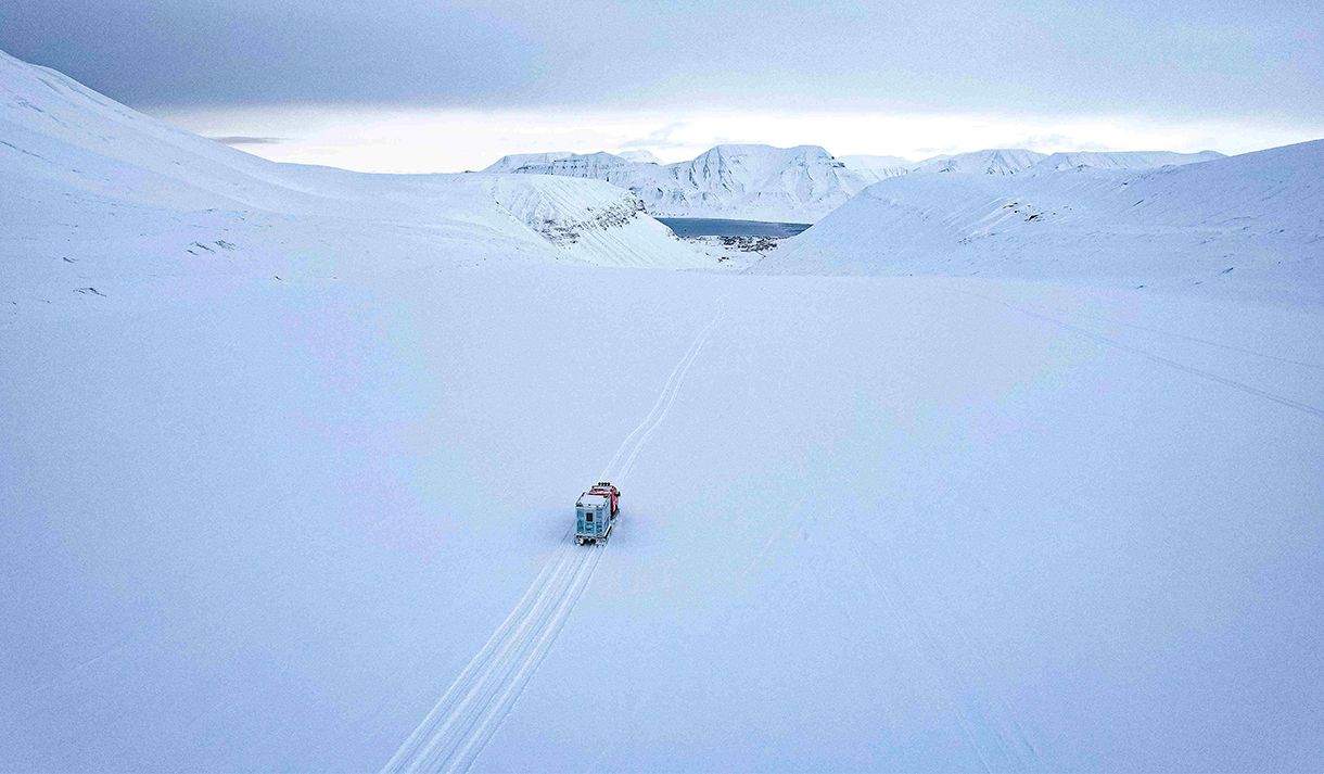 En beltevogn på en isbre som kjører langs spor i snøen til Longyearbyen i bakgrunnen