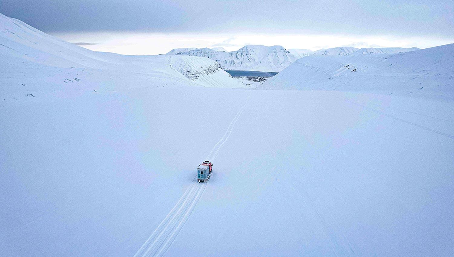 En beltevogn på en isbre som kjører langs spor i snøen til Longyearbyen i bakgrunnen