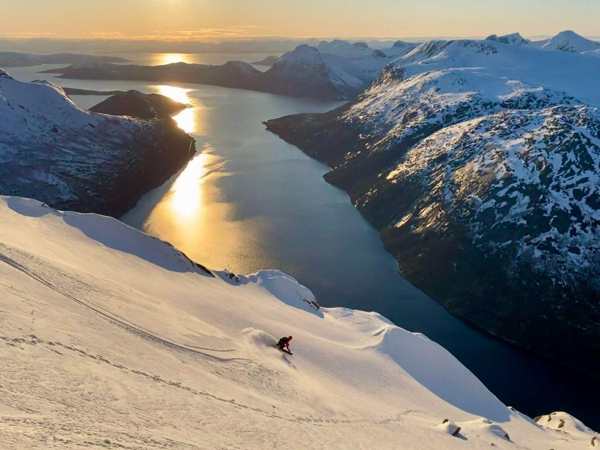 A lone skier descends a snowy mountainside above a long fjord, with golden sunset reflections on the water..