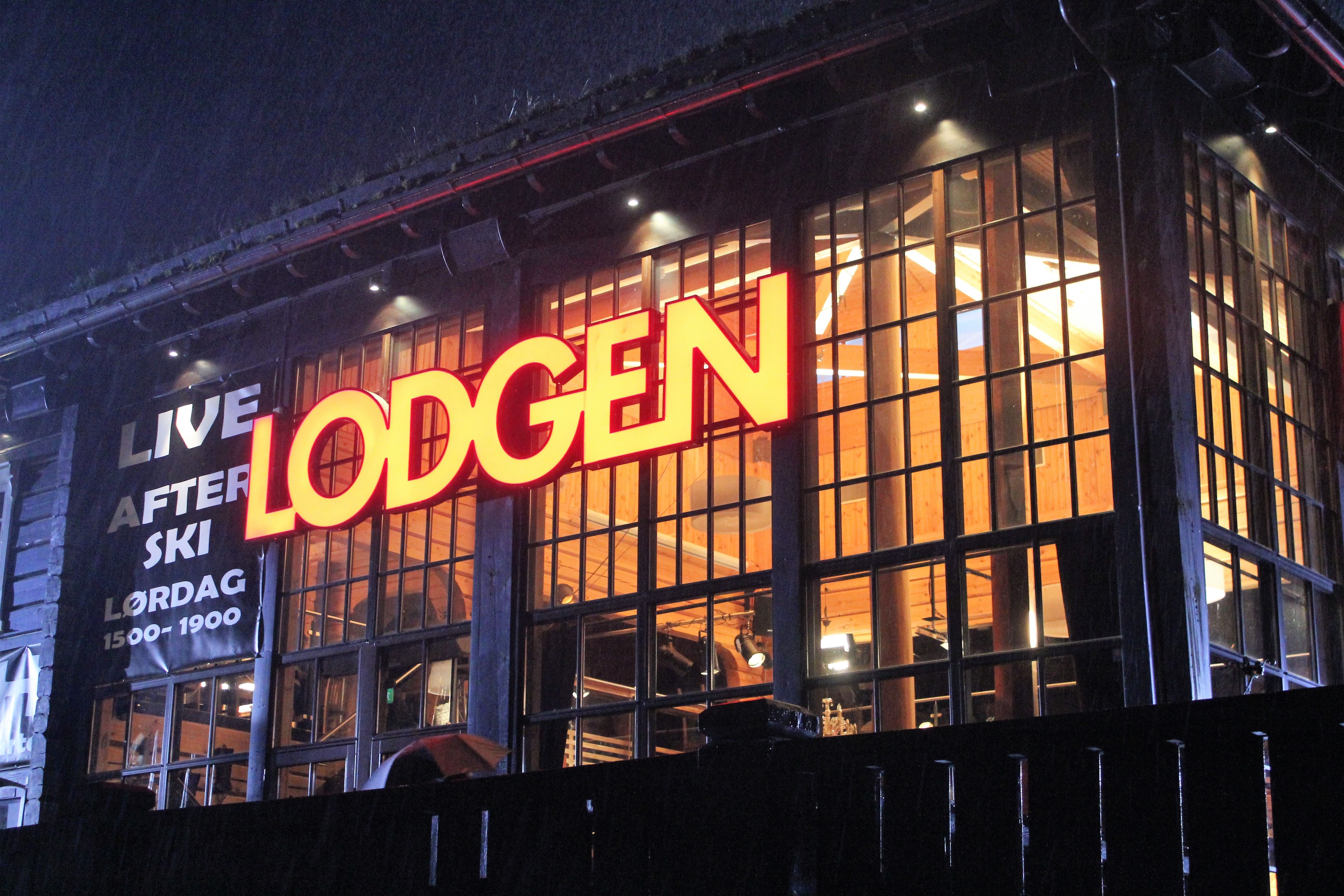 Kveldsbilde av bygning med store vinduer og rødt, opplyst skilt «LODGEN», med varmt lys inne..